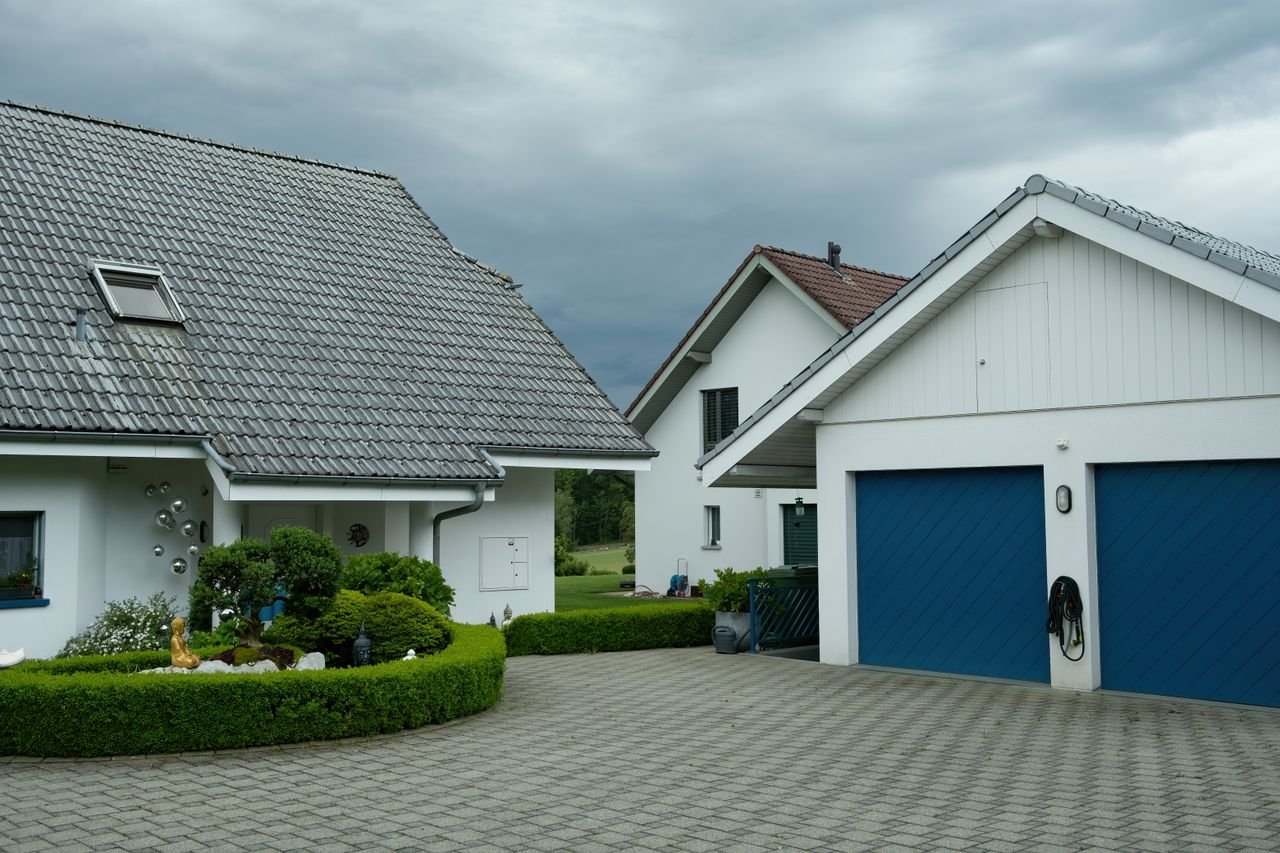 portfolio-01 Lovely suburban houses with blue garage doors under cloudy sky, surrounded by greenery.