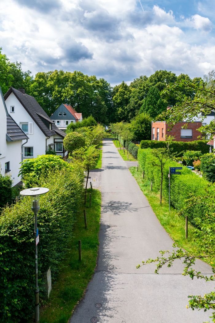 why-choose-us Peaceful suburban street lined with green hedges and houses under a partly cloudy sky.